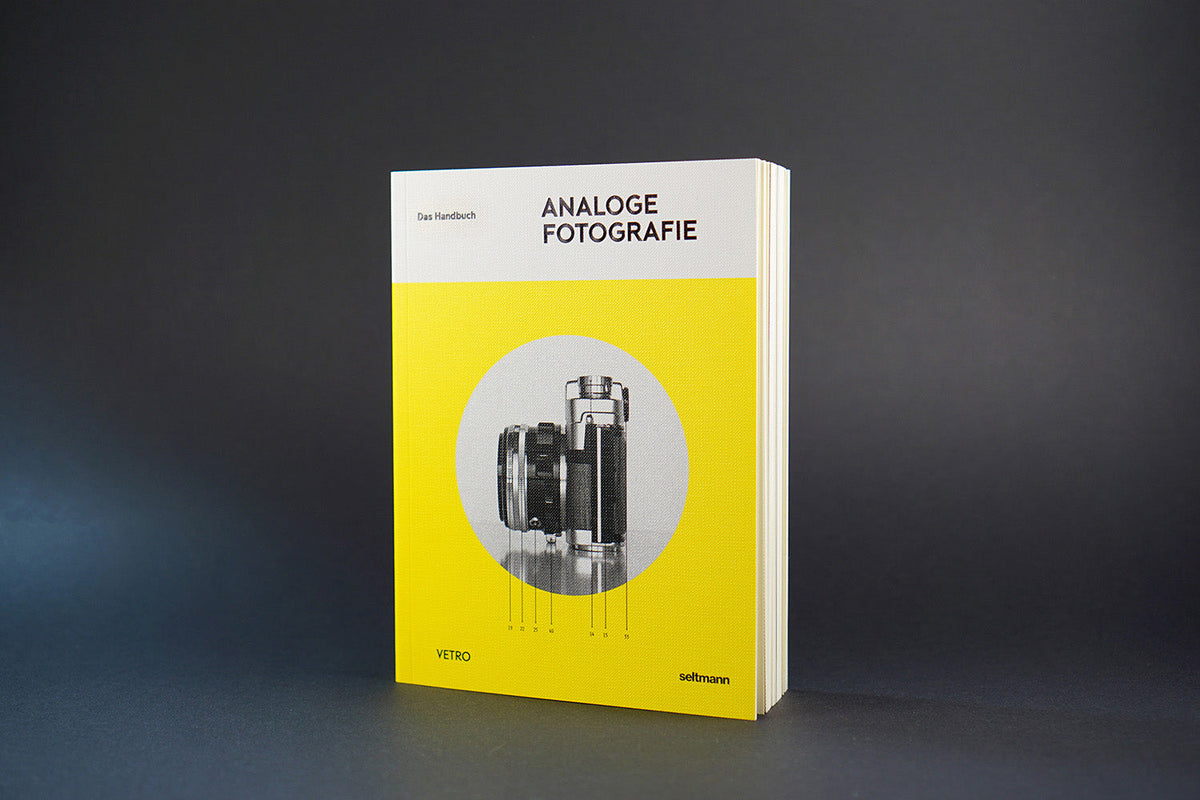 Aufrecht stehendes Buch mit gelbem Cover und Schwarz-Weiß-Foto einer analogen Kamera vor dunklem Hintergrund.