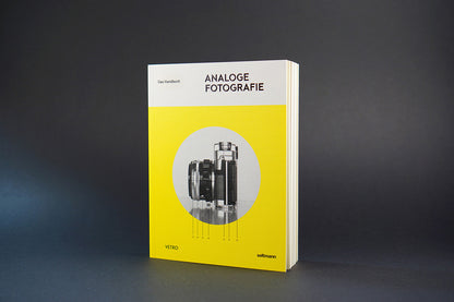 Aufrecht stehendes Buch mit gelbem Cover und Schwarz-Weiß-Foto einer analogen Kamera vor dunklem Hintergrund.