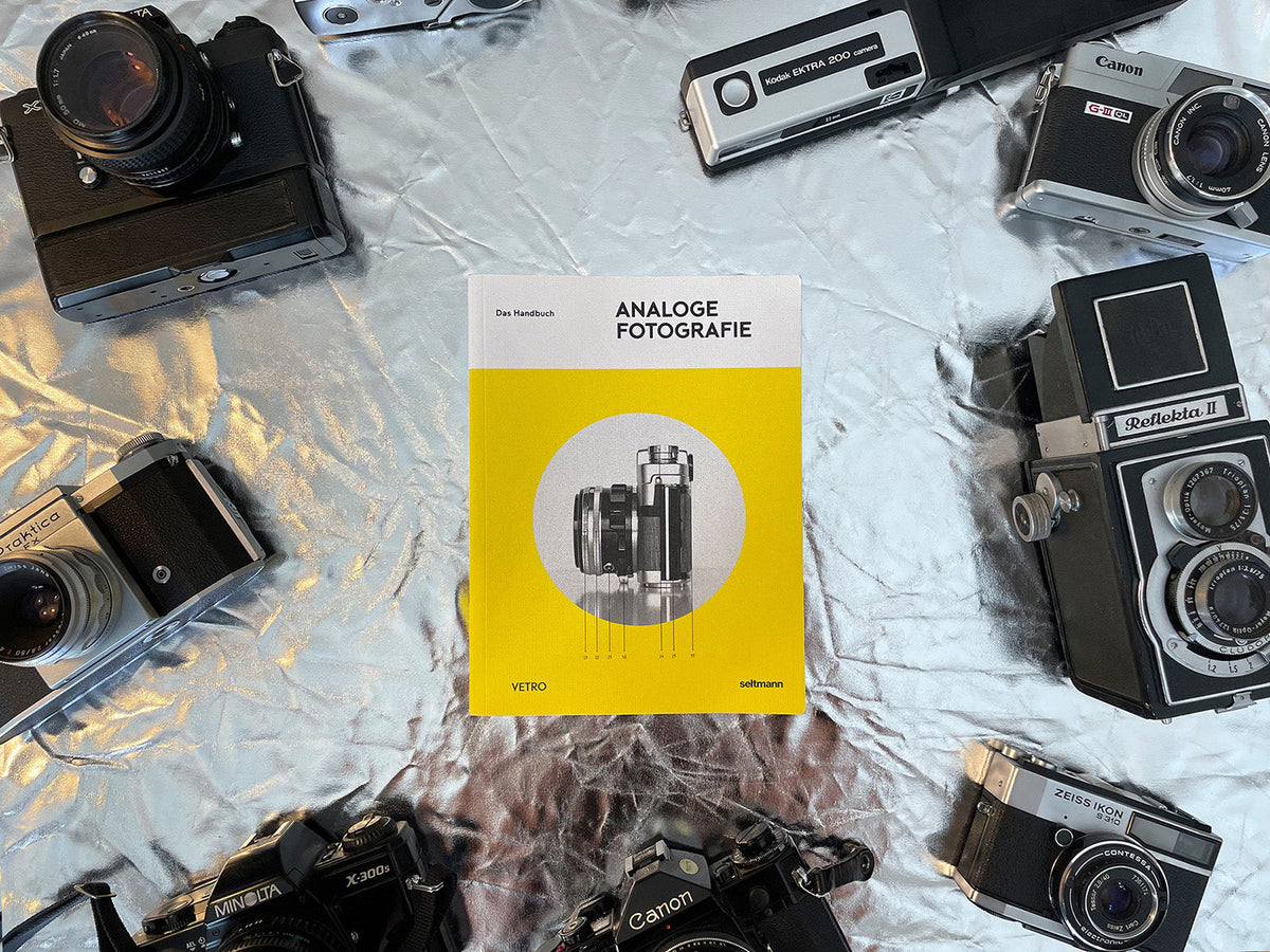 Das Buch Analog Fotografie von Seltmann Publishers liegt auf einer Folie zwischen mehreren analogen Kameras.