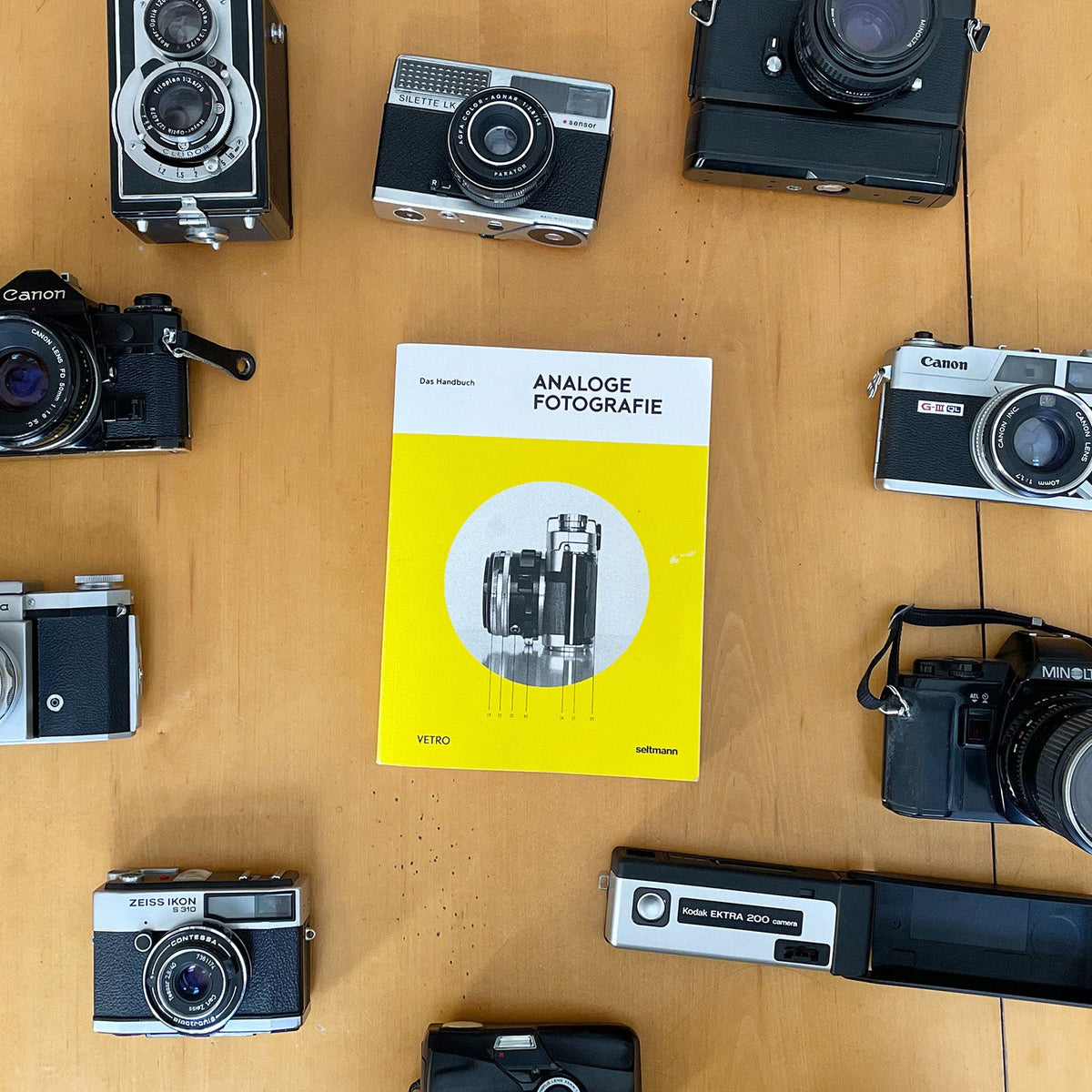 Das gelbe Handbuch Analoge Fotografie von Seltmann Publishers liegt auf einem Holztisch, umgeben von verschiedenen analogen Kameras.