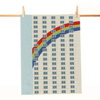 Geschirrtuch mit Motiv einer Hausfassade und Regenbogenstreifen an einer Holzleiste aufgehängt.