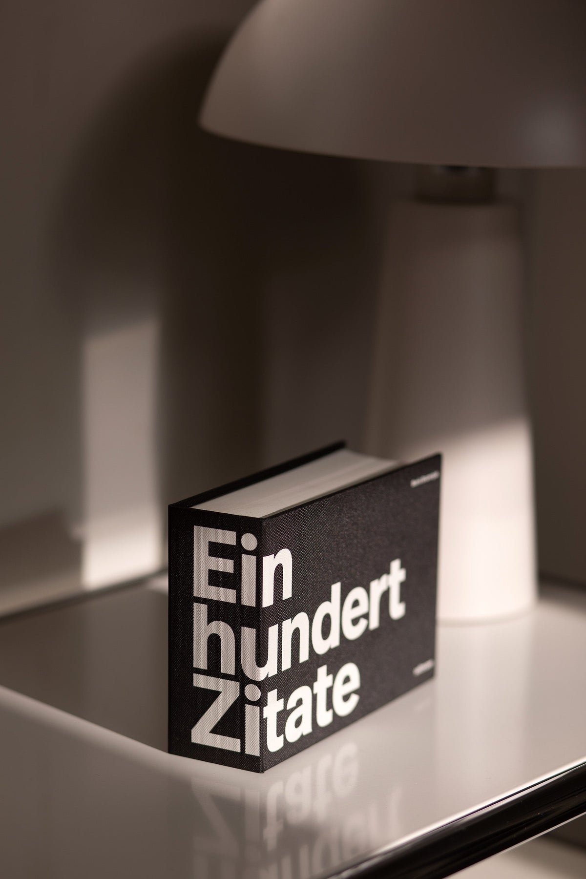 Das Buch Einhundert Zitate mit schwarzem Cover und weißer Typografie steht auf einer glänzenden Fläche neben einer Lampe.