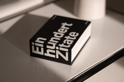 Das Buch Einhundert Zitate mit schwarzem Cover und weißer Typografie liegt auf einer hellen Tischplatte.