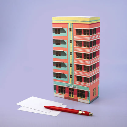 Hochformatiger Papierblock mit rosa Hotel-Fassade und Schriftzug HOTEL MIAMI neben Zetteln und rotem Stift.