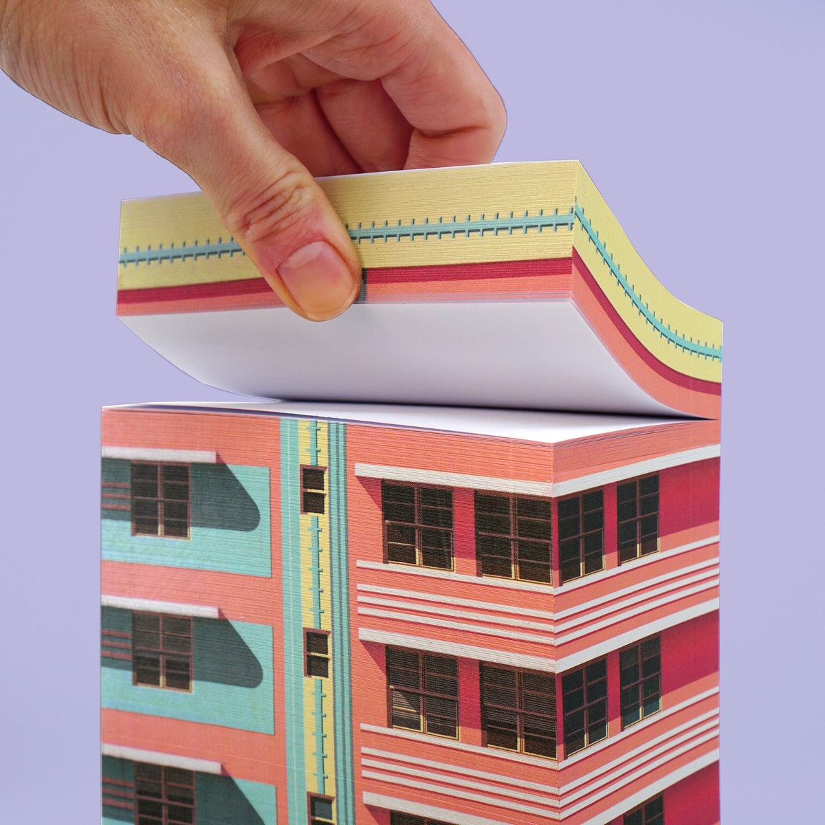 Hand hebt eine Seite eines Papierblocks mit Art-Deco-Gebäudemotiv in Rosa und Türkis vor violettem Hintergrund.
