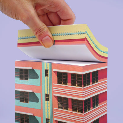 Hand hebt eine Seite eines Papierblocks mit Art-Deco-Gebäudemotiv in Rosa und Türkis vor violettem Hintergrund.