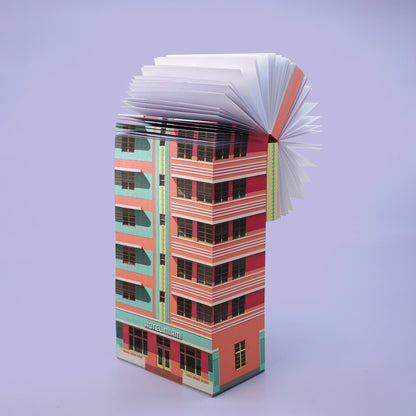 Aufgeschlagenes Buch mit gefalteten Seiten, die ein rosa-türkises Hochhaus mit der Aufschrift Hotel miami darstellen.