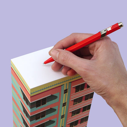 Hand schreibt mit rotem Stift auf einen Notizblock, dessen Seitenflächen ein Motiv eines rosa-grünen Hochhauses zeigen.