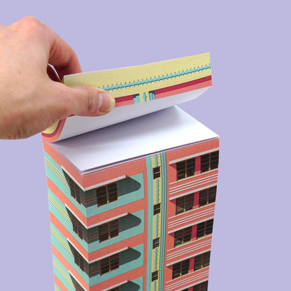 Eine Hand reißt ein Blatt von einem Papierblock mit Motiv eines rosa-türkisen Hochhauses vor lila Hintergrund.